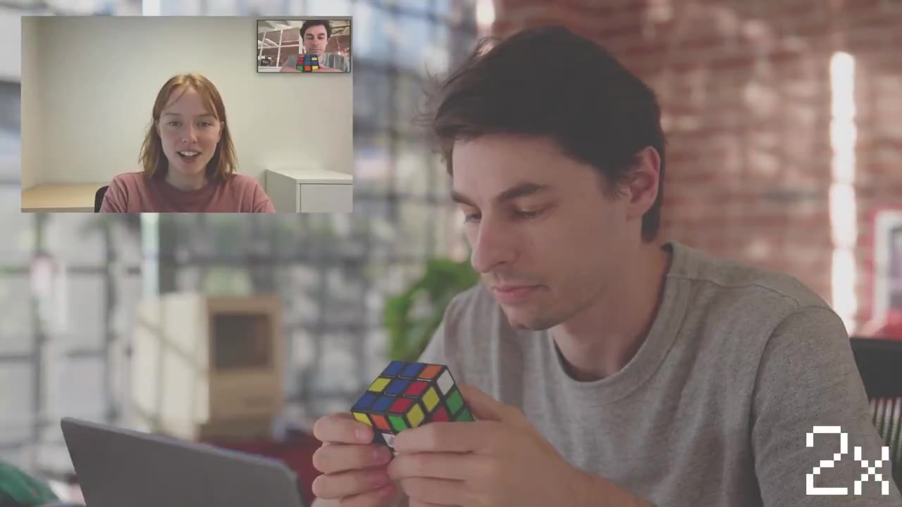 Een man draait aan een Rubiks kubus terwijl een AI-persona in een videovenster meekijkt, rechtsonder staat 2x